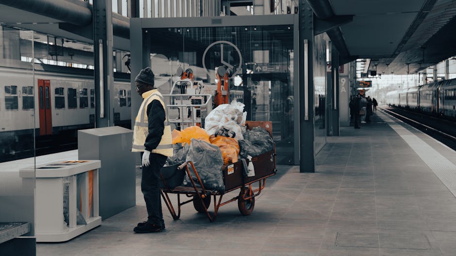 A worker dressed in dark clothing, wearing a grey beanie and a high-visibility vest, stands on a train station platform beside a large red wheeled trolley loaded with various waste bags and debris. Th