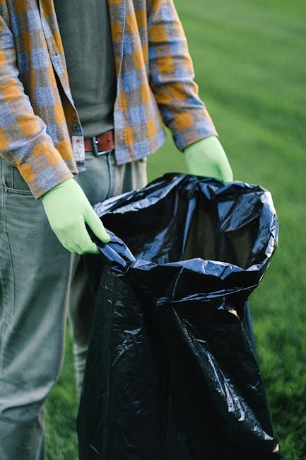 A worker dressed in dark clothing, wearing a grey beanie and a high-visibility vest, stands on a train station platform beside a large red wheeled trolley loaded with various waste bags and debris. Th