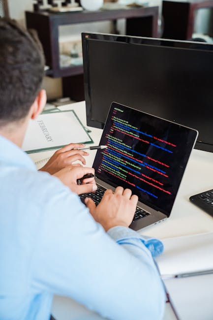 A person wearing a light blue shirt is seated at a white desk, working on a silver laptop displaying lines of colorful code on the screen. The individual is using a black mouse with their right hand w